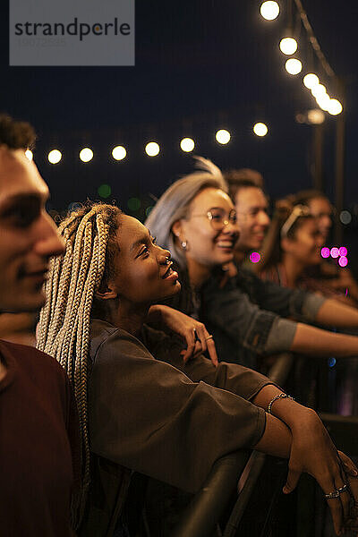 Happy friends having social gathering under string lights on rooftop