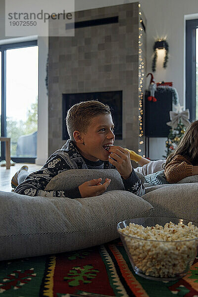 Happy boy eating popcorn by sister and watching TV at home