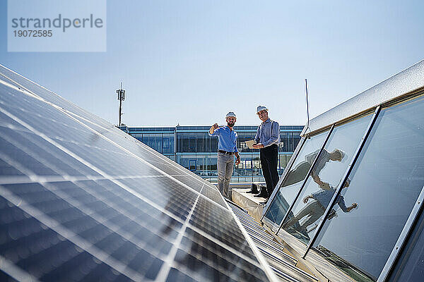 Zwei Geschäftsleute mit Schutzhelmen treffen sich auf dem Dach eines Firmengebäudes mit Solarpaneelen