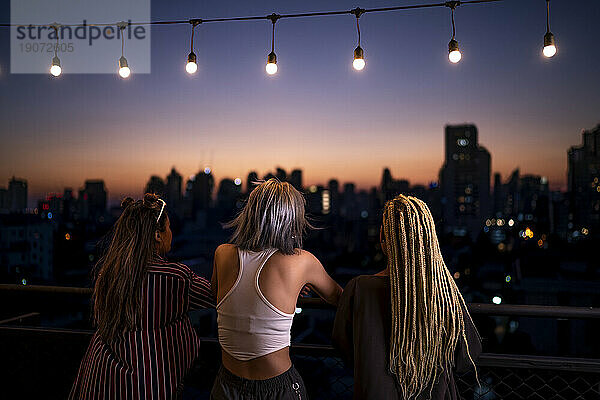 Freunde stehen in der Abenddämmerung unter Lichterketten auf dem Dach