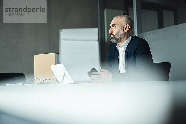 Nachdenklicher reifer Geschäftsmann sitzt mit Smartphone am Schreibtisch