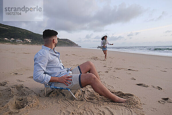 Woman running towards boyfriend sitting on chair at beach