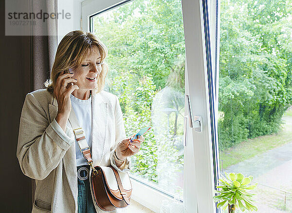 Reife Frau mit Kreditkarte telefoniert am Fenster