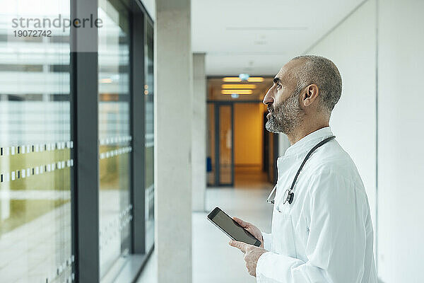 Nachdenklicher reifer Arzt mit Tablet-PC  der durch das Fenster im Krankenhaus schaut