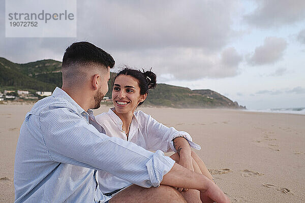 Glückliche junge Frau schaut ihrem Freund zu  der am Strand sitzt