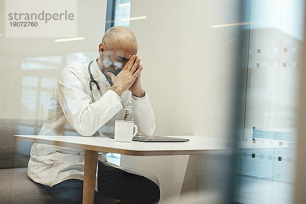 Müder reifer Arzt sitzt mit Kaffeetasse und Laptop durch Glas gesehen