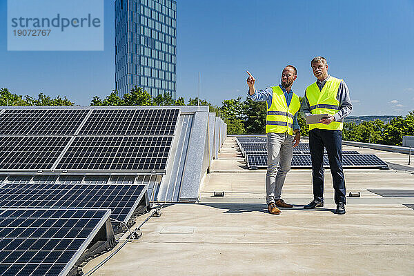 Zwei Männer mit Tablet-PC und reflektierenden Westen unterhalten sich auf dem Dach eines Firmengebäudes mit Solarpaneelen