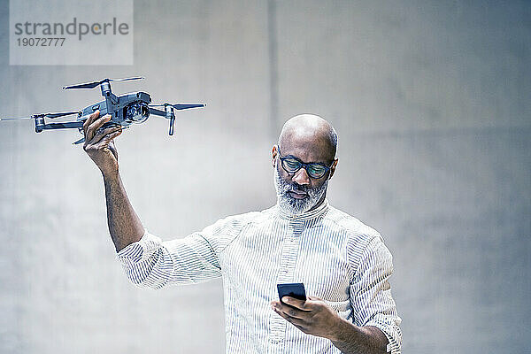 Porträt eines glatzköpfigen reifen Mannes  der einen Quadcopter in der Hand hält und dabei auf sein Handy schaut