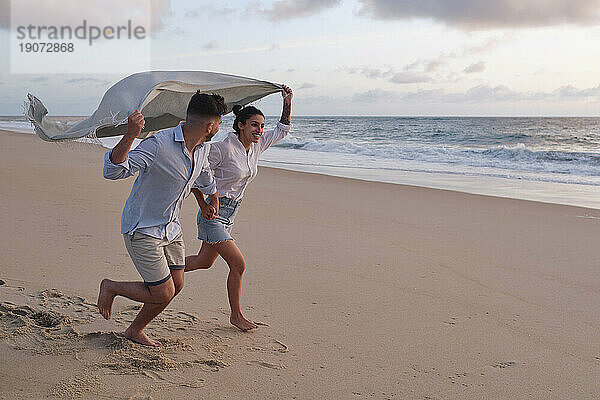 Fröhliches Paar läuft zusammen und hält eine Decke am Strand