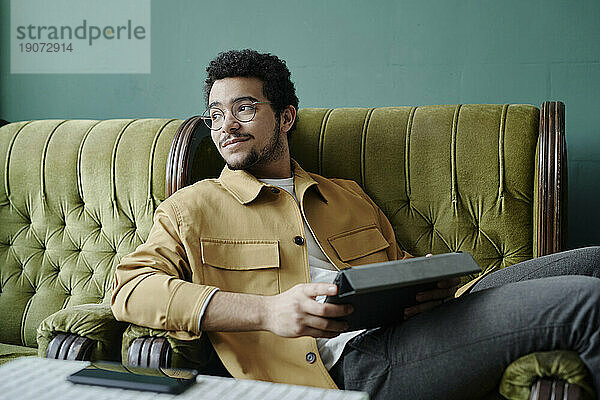Nachdenklicher junger Geschäftsmann sitzt entspannt im grünen Sessel und hält einen Tablet-PC in der Hand