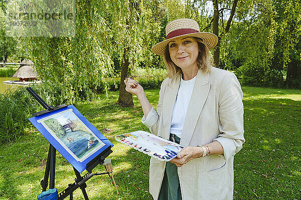 Lächelnde reife Frau steht mit Aquarellen im Park