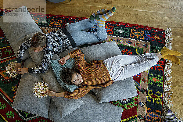 Siblings with popcorn bowls lying on pillows at home
