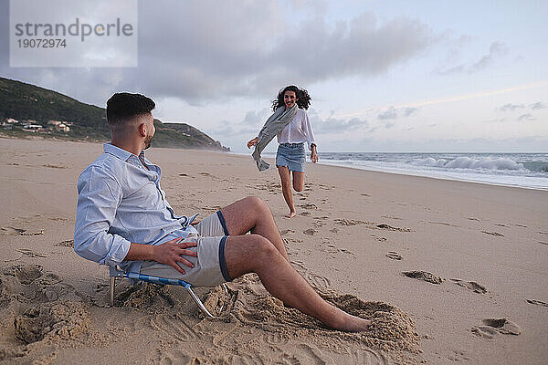 Man sitting on chair looking at girlfriend running on beach