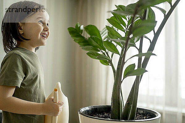 Lächelnder Junge hält Gießkanne neben Pflanzen zu Hause