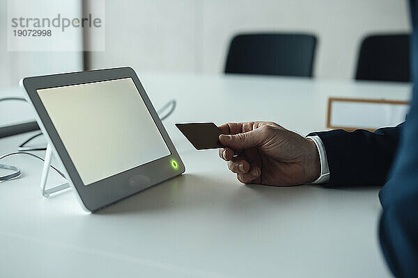 Reifer Geschäftsmann mit Kreditkarte und Tablet-PC sitzt am Schreibtisch