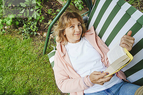 Happy mature woman lying on swing with book