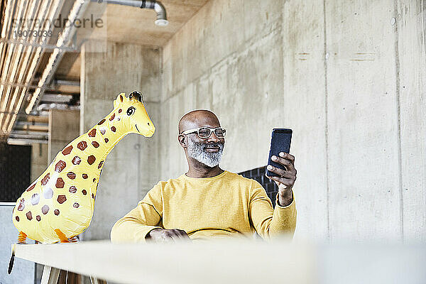 Lächelnder reifer Geschäftsmann sitzt am Schreibtisch im Büro mit Handy und Giraffenfigur
