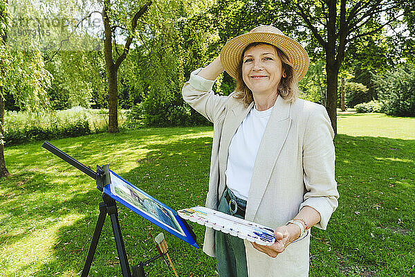 Lächelnde reife Frau mit Hut steht mit Aquarellfarben im Park