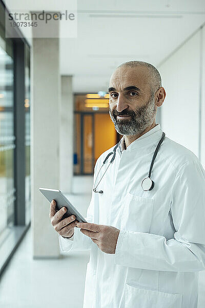 Lächelnder reifer Arzt steht mit Tablet-PC im Krankenhaus