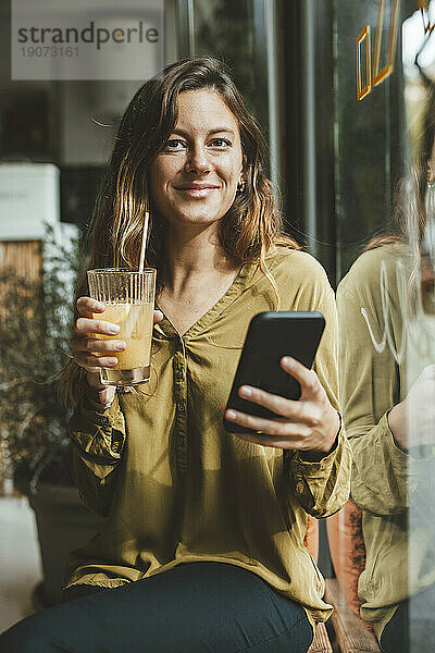 Lächelnde Frau sitzt mit einem Glas Saft und einem Smartphone im Café