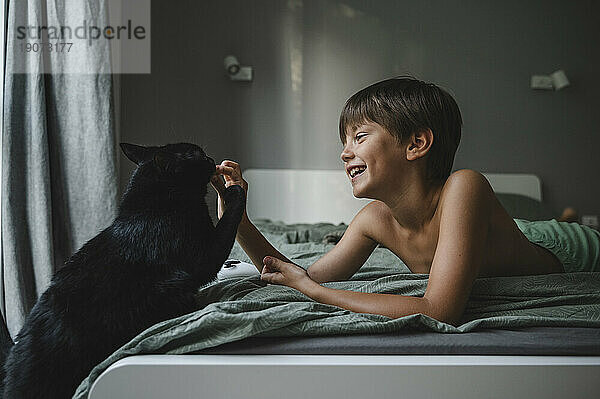 Lächelnder Junge liegt auf dem Bett und spielt zu Hause mit der schwarzen Katze