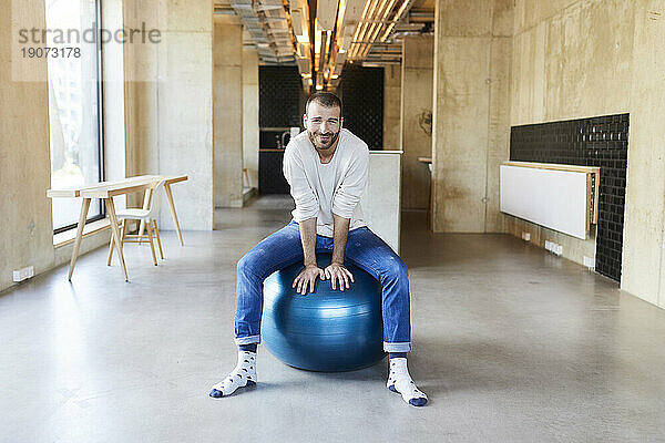 Porträt eines jungen Mannes  der in einem modernen Büro auf einem Fitnessball sitzt