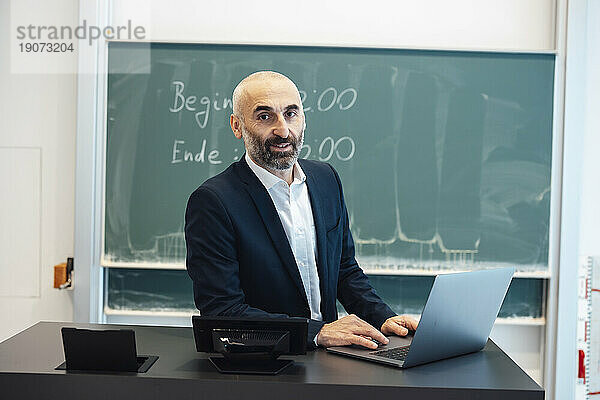 Professor sitzt mit Laptop am Schreibtisch im Hörsaal