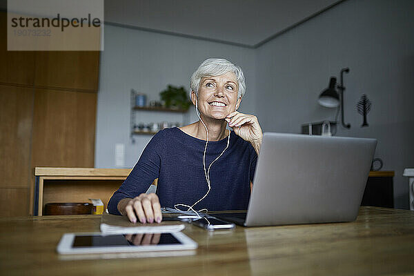 Aktive ältere Frau hört Musik auf dem Smartphone und benutzt einen Laptop  während sie zu Hause wegschaut