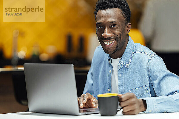 Glücklicher Geschäftsmann hält Tasse mit Laptop im modernen Büro