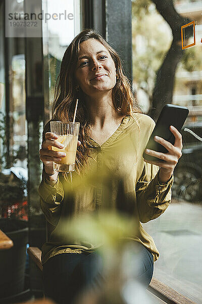 Lächelnde junge Frau hält ein Glas Saft und ein Smartphone im Café