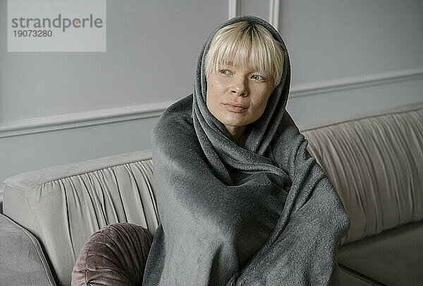 Contemplative woman wrapped in blanket sitting on sofa at home