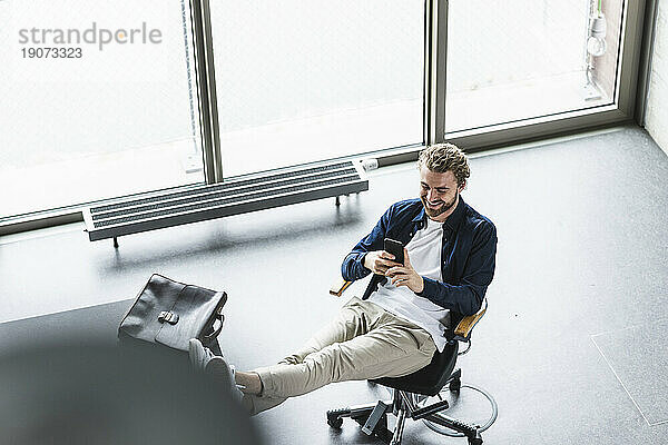 Lächelnder  lässiger Geschäftsmann  der mit erhobenen Füßen im Büro sitzt und sein Handy benutzt