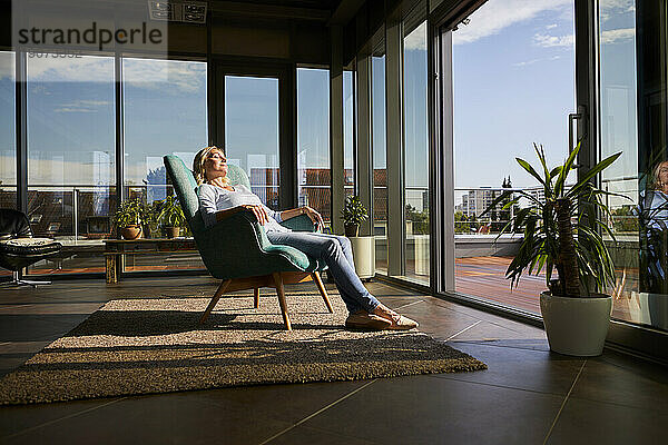 Reife Frau entspannt sich zu Hause im Sessel im Sonnenlicht