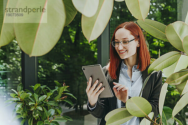 Lächelnde Geschäftsfrau nutzt Tablet-PC bei Pflanzen im Büro