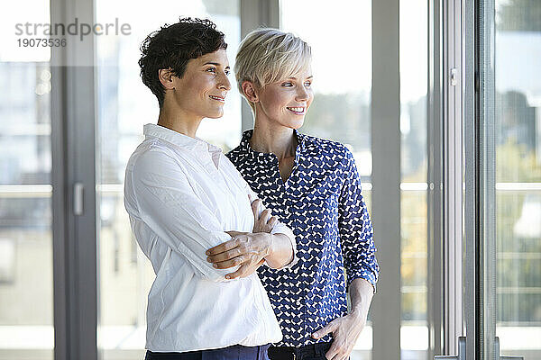 Zwei lächelnde Geschäftsfrauen schauen aus dem Fenster im Büro