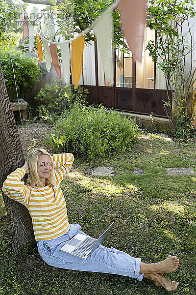 Lächelnder Freiberufler mit Laptop  der unter einem Baum im Garten ruht