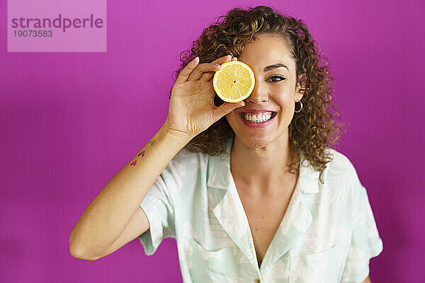 Glückliche Frau hält eine Zitronenscheibe über dem Auge vor magentafarbenem Hintergrund