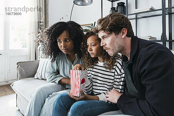 Eltern und Tochter sitzen auf dem Sofa und essen Popcorn und schauen sich einen Film auf dem Laptop an