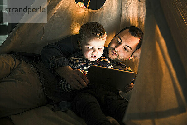 Father and son sharing a tablet in a dark tent at home