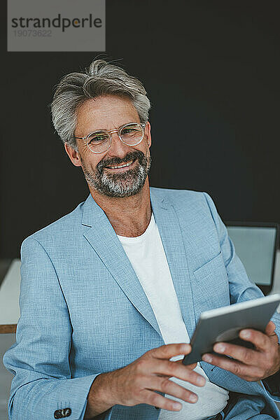 Lächelnder Geschäftsmann mit Tablet-PC sitzt im Büro