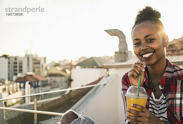 Porträt einer glücklichen jungen Frau  die bei Sonnenuntergang auf dem Dach etwas trinkt