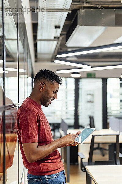 Lächelnder Geschäftsmann mit Tablet-PC  der sich im Büro lehnt