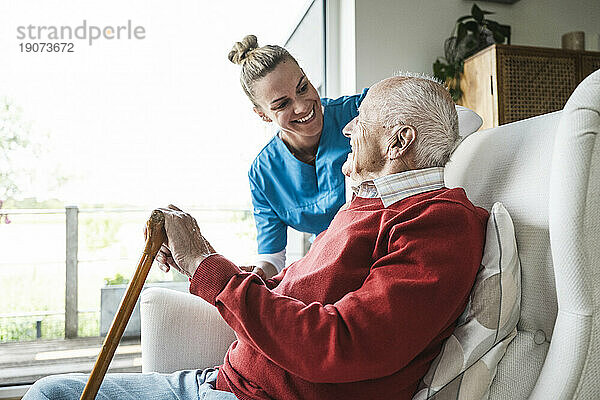 Glückliche Krankenschwester  die sich um den älteren Mann kümmert  der zu Hause im Sessel sitzt