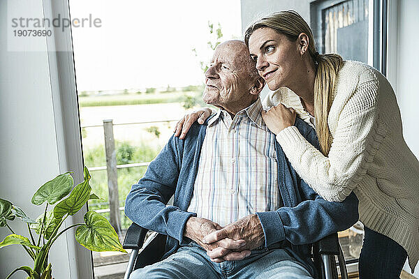Lächelnde blonde Frau kümmert sich um den Vater  der zu Hause im Rollstuhl sitzt