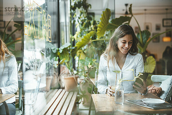 Lächelnde junge Frau benutzt Smartphone am Tisch im Café