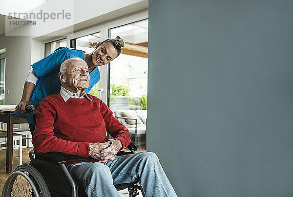 Glückliche Krankenschwester  die sich um einen älteren Mann kümmert  der zu Hause im Rollstuhl an der Wand sitzt