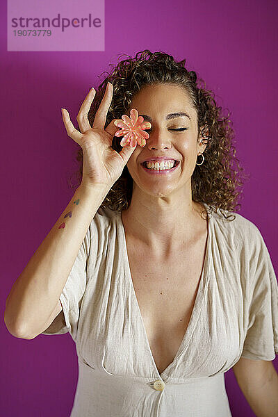 Fröhliche Frau bedeckt Auge mit Blume vor magentafarbenem Hintergrund
