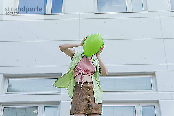 Frau bedeckt Gesicht mit grünem Luftballon vor Gebäude