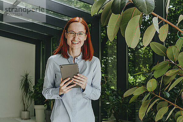 Fröhliche rothaarige Geschäftsfrau steht mit Tablet-PC neben der Fabrik im Büro