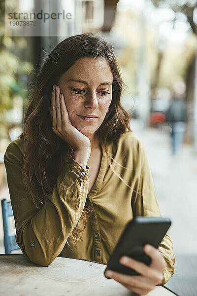 Junge Frau mit der Hand am Kinn benutzt Smartphone im Café